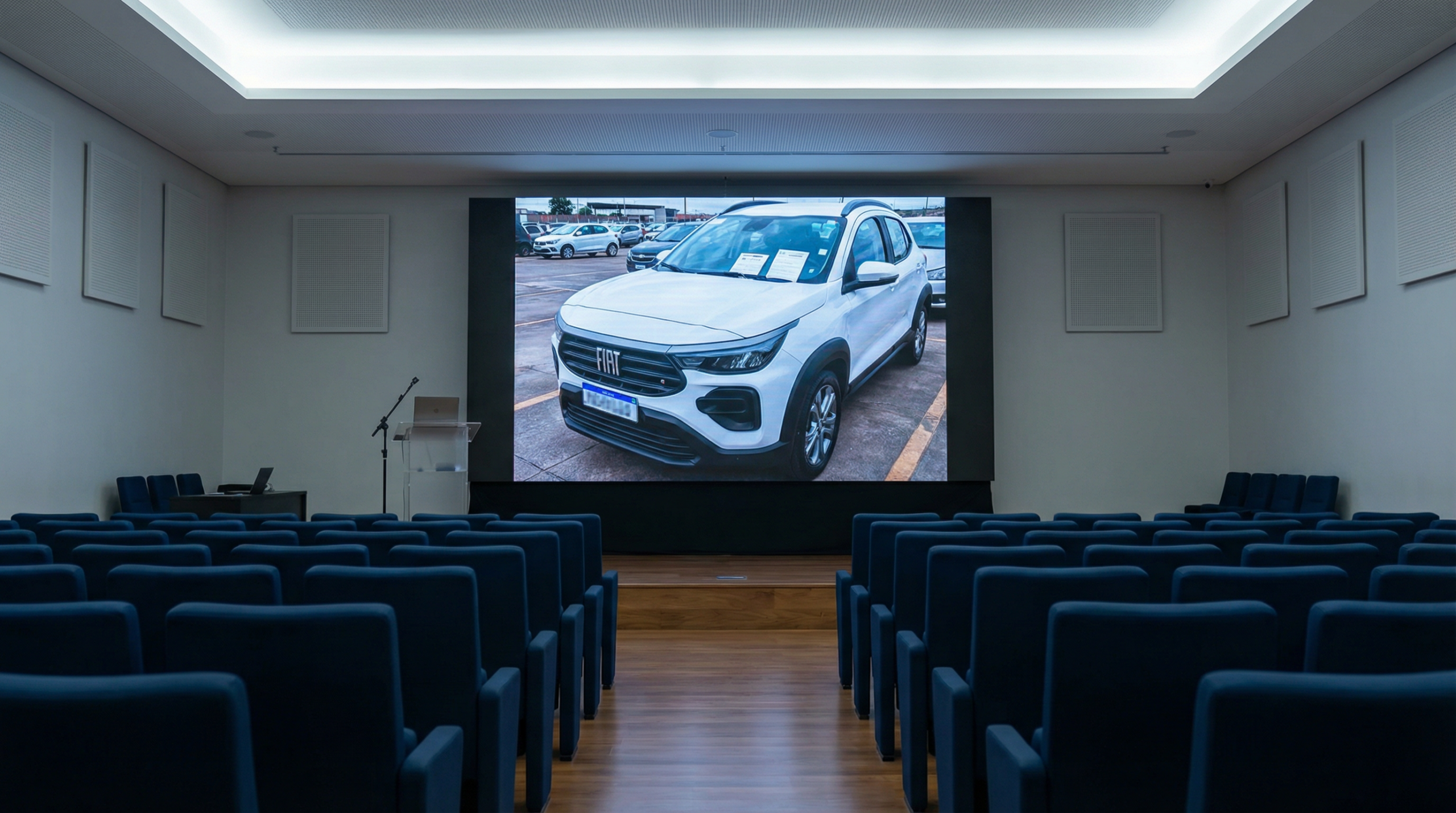 Salão de leilão presencial com cadeiras, telão mostrando um SUV branco, e ambiente formal.